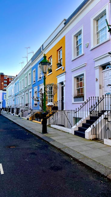 A narrow cobblestone street in a residential area of West Kensington, lined with multi-story terraced houses featuring small front gardens with potted plants and flower beds. Some houses have bay wind