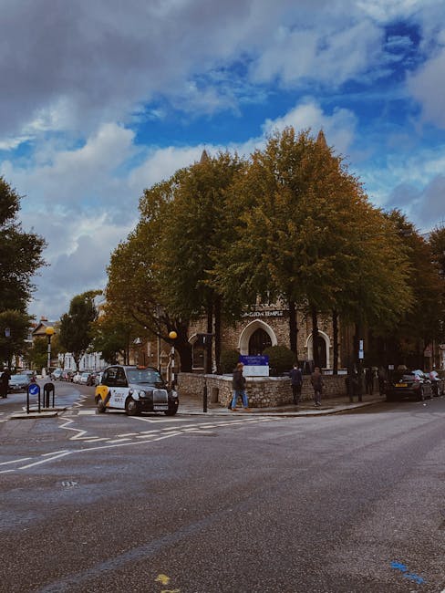 A street scene outside a historic brick building with an arched entrance and a sign that reads 'Fulham Palace'. Several large deciduous trees with green and brown foliage partially obscure the buildin